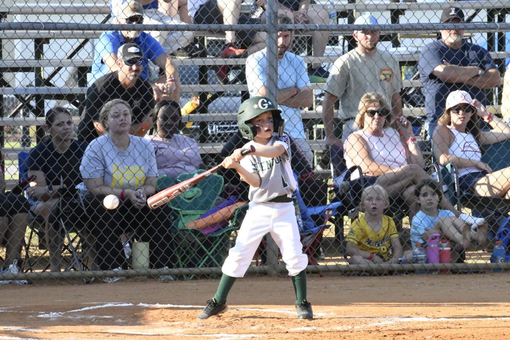 A young child in baseball gear stands at bat, swinging at a baseball during a game, with spectators watching from bleachers behind a fence.