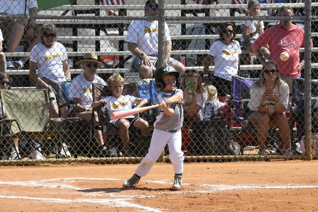 Child swinging a bat during a baseball game, wearing helmet and uniform, with spectators sitting in bleachers behind a chain-link fence.