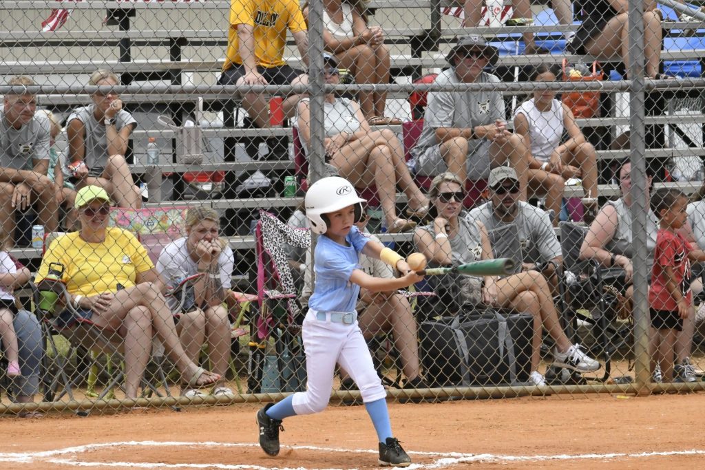 A young child wearing a helmet swings a bat at a baseball game, with spectators sitting in bleachers behind a chain-link fence.