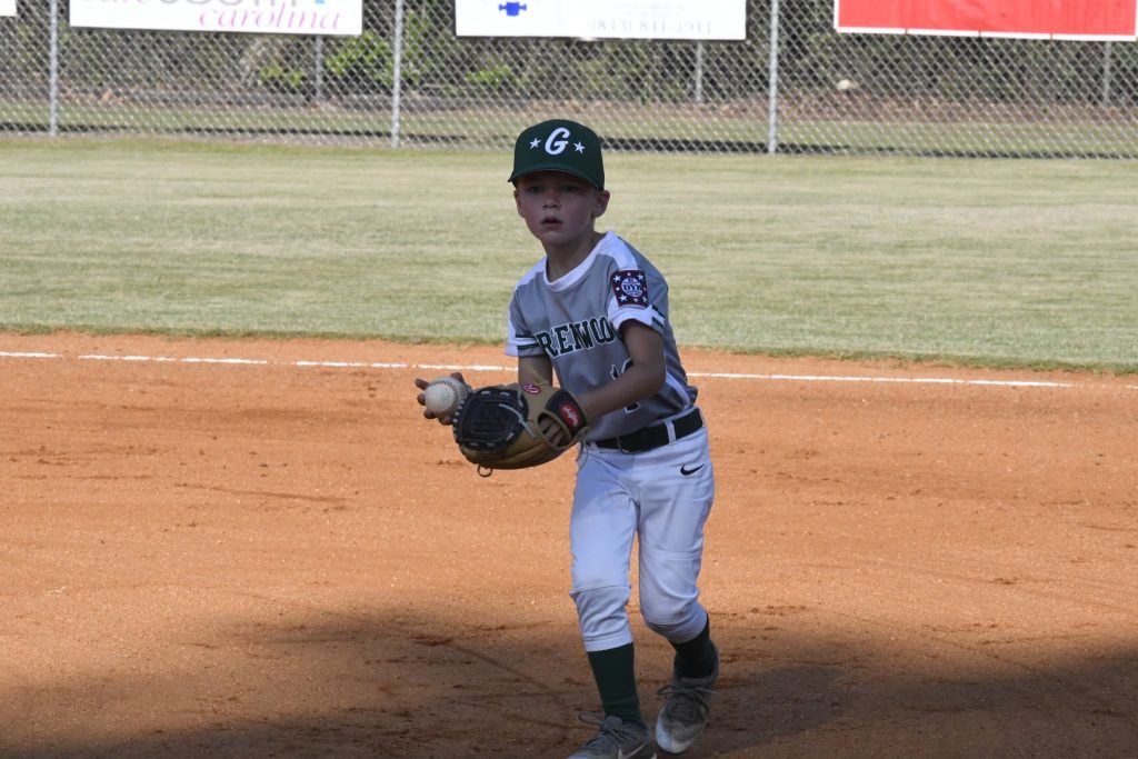 A young boy in a baseball uniform stands on a dirt field, wearing a glove and cap, ready to catch a ball during a game.