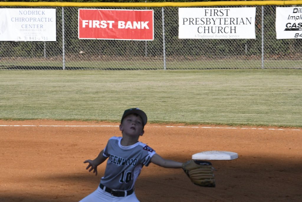 A young baseball player reaches up to catch a ball on a sunny field, with sponsorship signs visible on the fence in the background.