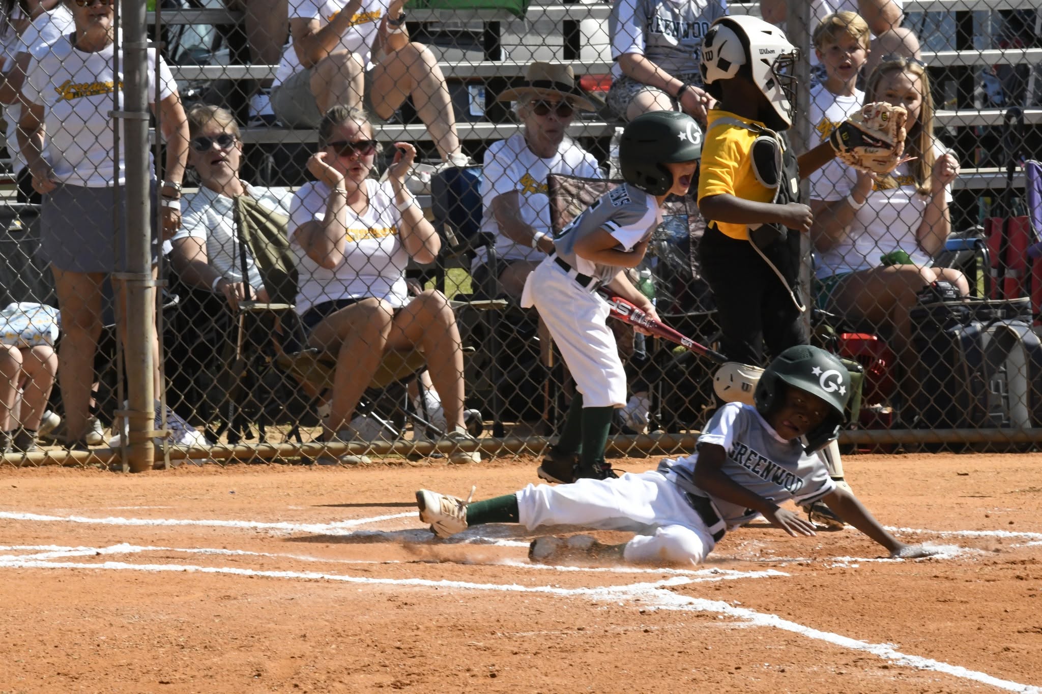 A young baseball player slides into home plate as spectators and teammates watch and cheer from the stands.