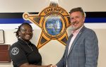 A sheriff’s deputy and a man in a suit shake hands, standing in front of a large sheriff’s emblem on the wall.