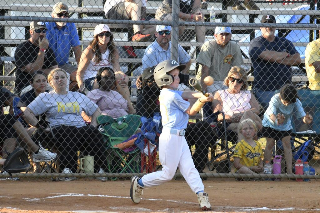 A young baseball player swings a bat during a game, with spectators seated in bleachers behind a chain-link fence watching attentively.