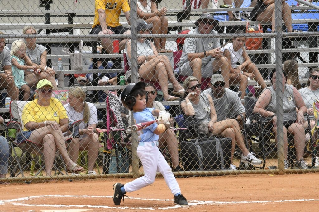 A young child in a baseball uniform swings a bat during a game, while spectators watch from bleachers behind a chain-link fence.