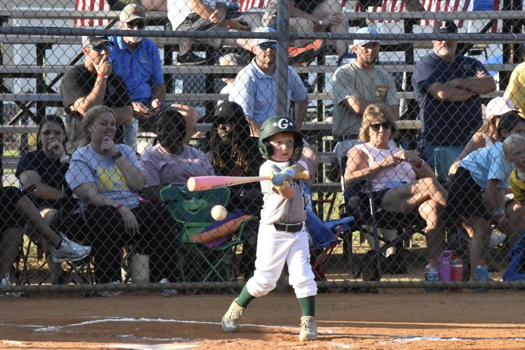 A young child in a baseball uniform swings a bat during a game, with spectators seated in bleachers watching behind a chain-link fence.