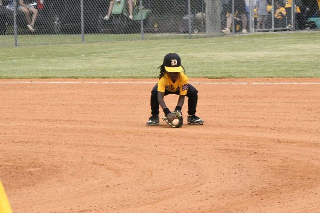 A young baseball player in a yellow jersey and black cap fields a ground ball on a dirt infield during a game.