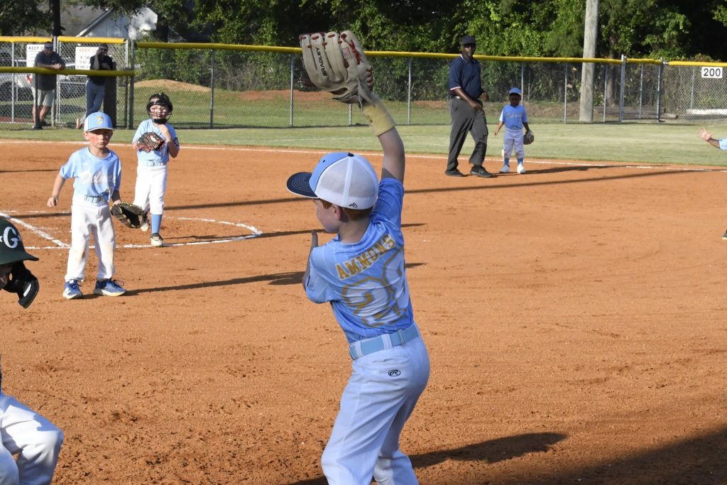 Young boy in a blue baseball uniform throwing a ball on a baseball field during a youth game, with other children and an umpire in the background.