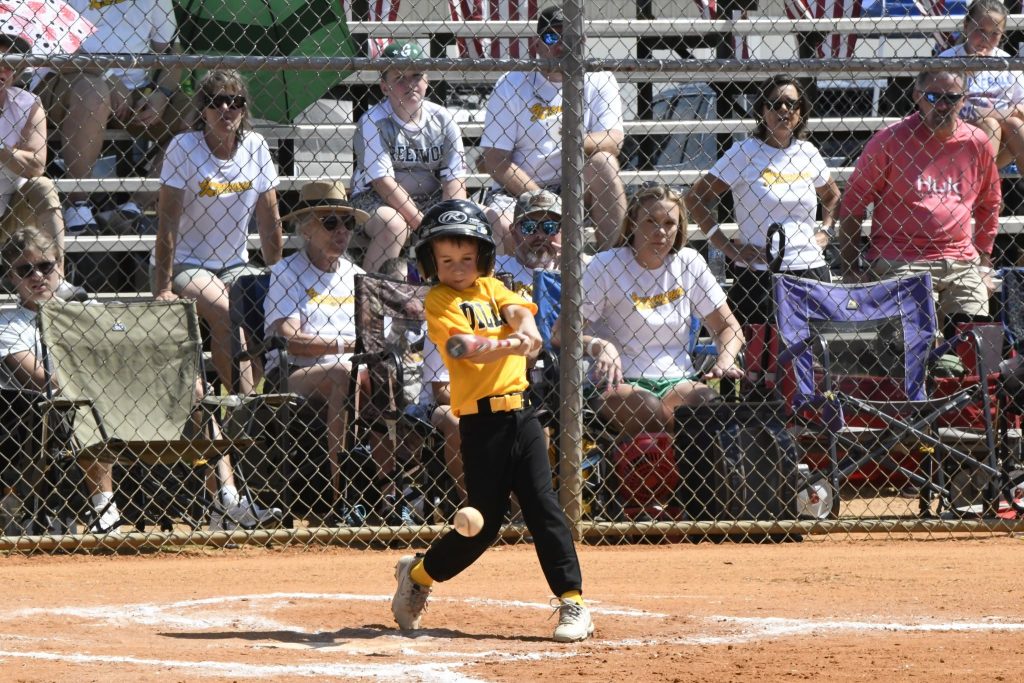 A young baseball player in a yellow jersey swings a bat during a game as spectators watch from the bleachers behind a chain-link fence.