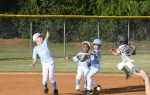 Four young kids playing baseball, two in blue uniforms reaching for the ball, while another player runs toward base on a sunny day.