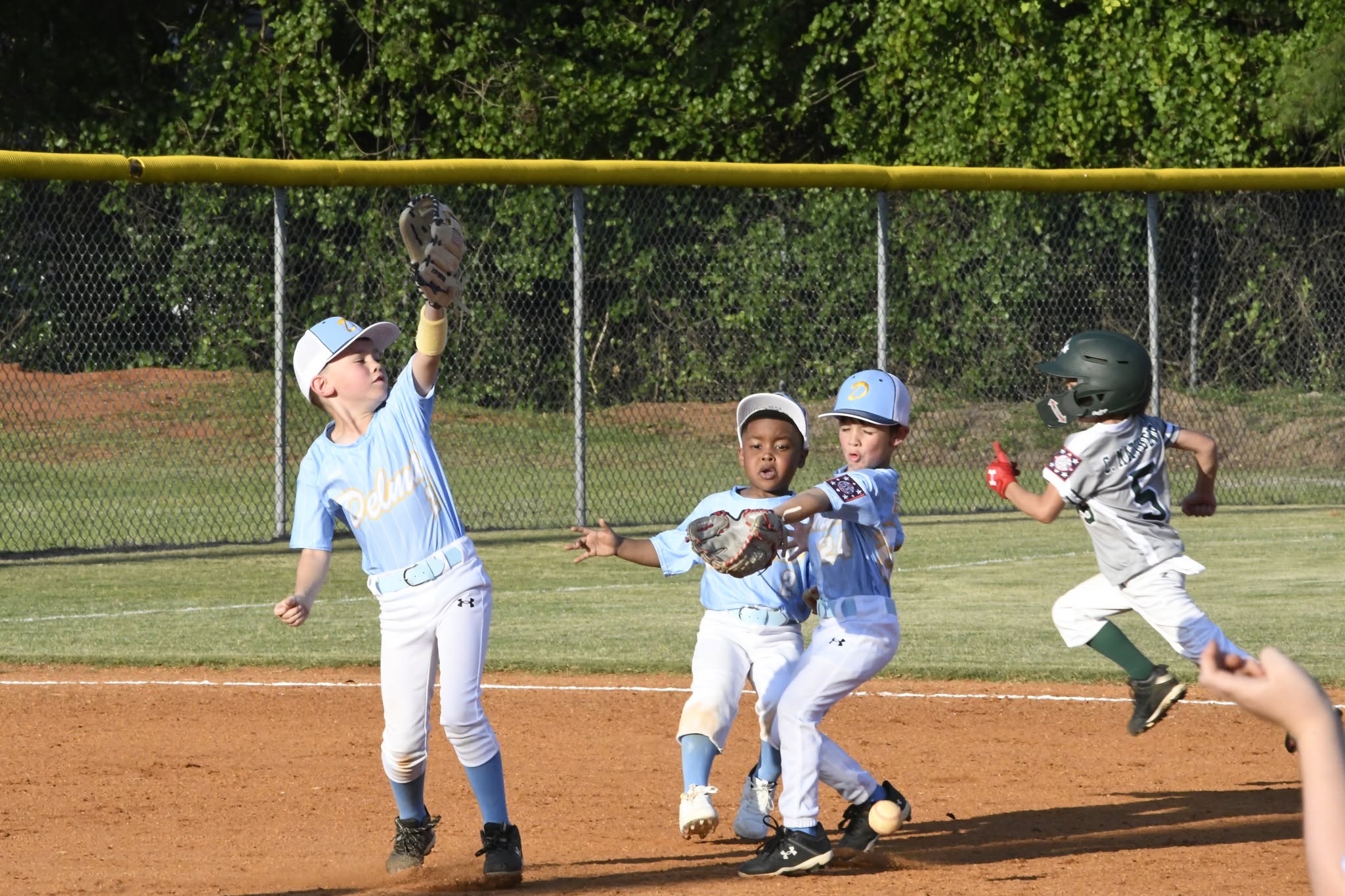Four young kids playing baseball, two in blue uniforms reaching for the ball, while another player runs toward base on a sunny day.