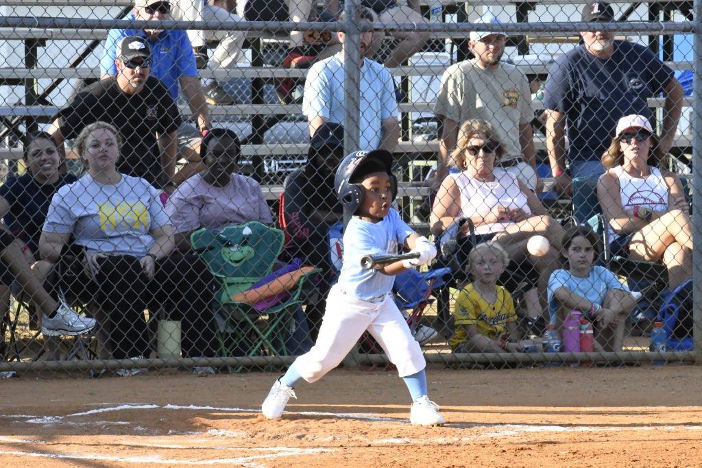 A young child in a baseball uniform swings a bat at home plate, with spectators watching from bleachers behind a fence.