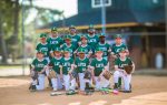 Youth baseball team in green uniforms posing on a baseball field with coaches, bats, and gloves on the ground in front of them.