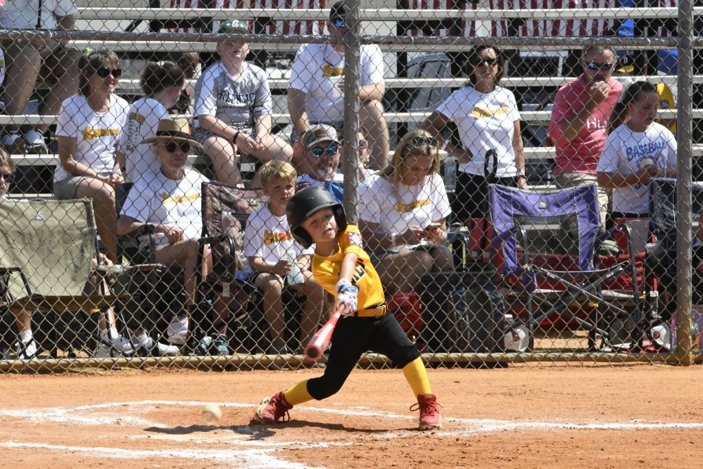 A young child in a yellow uniform swings a bat during a baseball game, with spectators watching from bleachers behind a chain-link fence.