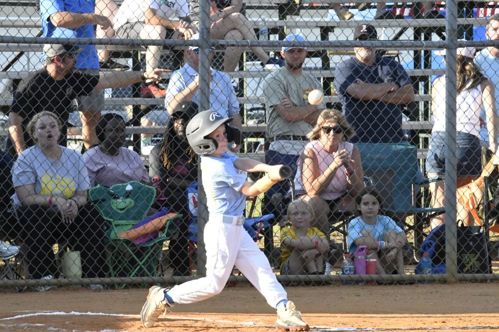 A young baseball player swings a bat during a game as spectators watch from the bleachers behind a chain-link fence.