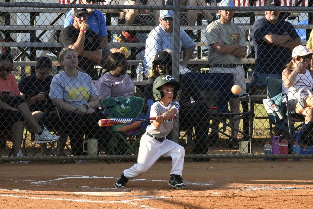 A young child swings a bat at a baseball during a game, with spectators watching from bleachers behind a chain-link fence.