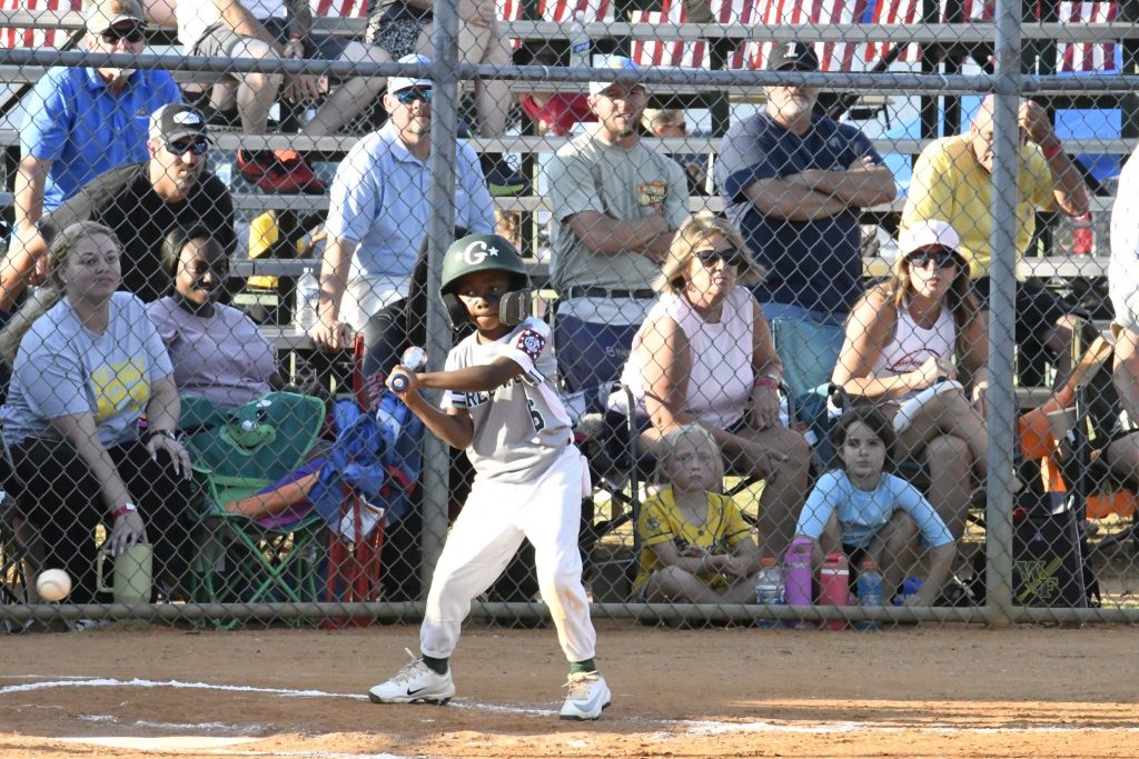 A young baseball player at bat prepares to swing as spectators watch from behind a chain-link fence at a sunny outdoor game.