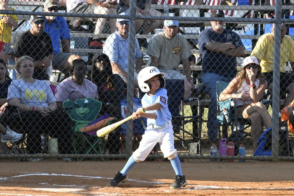 Young child in baseball uniform swings bat during a game, with spectators watching from the bleachers behind a chain-link fence.