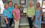 Five women stand indoors near a shelf with flowers and a framed certificate, smiling at the camera.