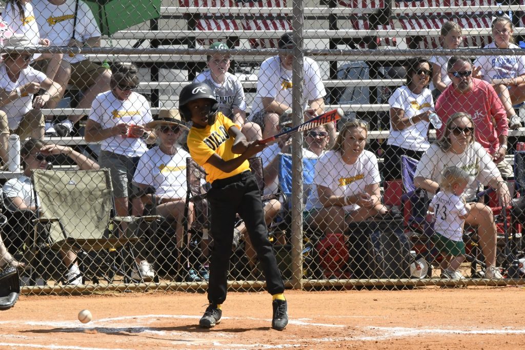 A young baseball player in a yellow jersey swings a bat during a game while spectators watch from bleachers behind a chain-link fence.