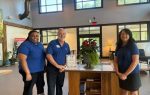 Three people wearing blue shirts stand smiling behind a reception desk in a bright, modern lobby with large windows and indoor plants.