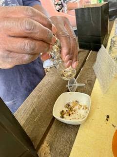 A person’s hands holding glassware and adding dried ingredients into a small white bowl on a wooden table.