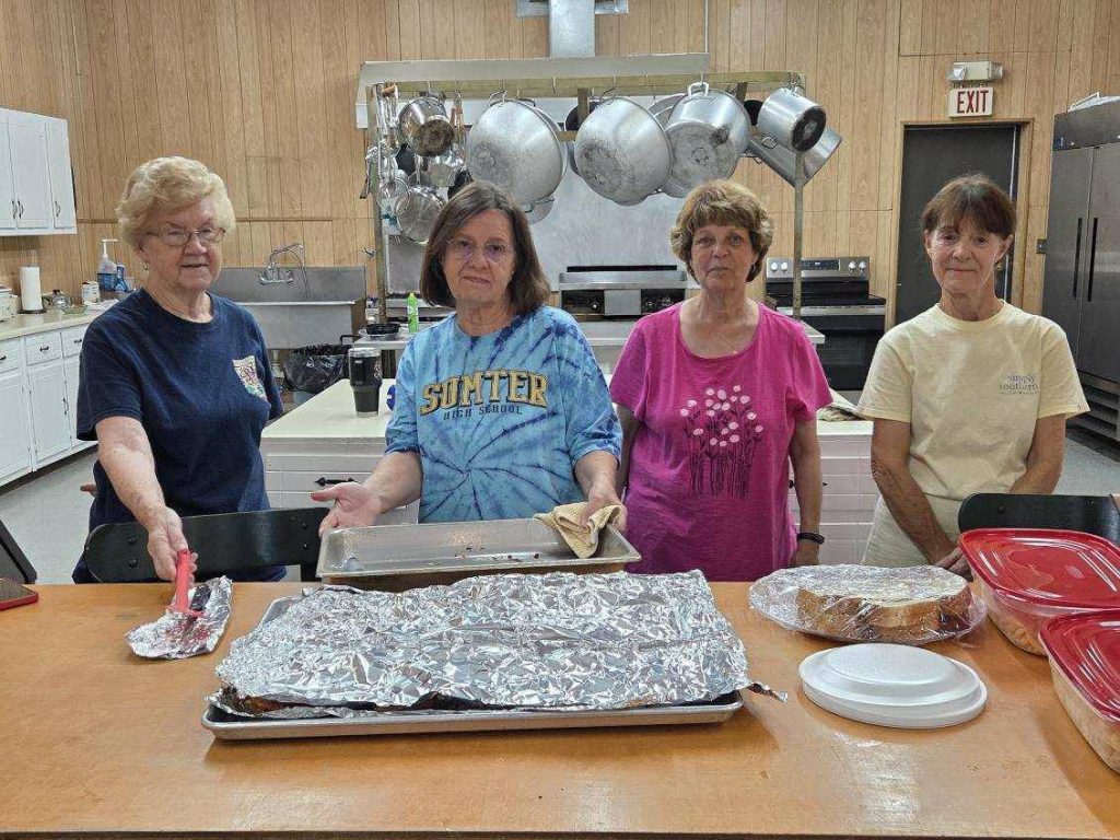 Four women stand behind a kitchen counter with covered trays and food containers, appearing to prepare or serve food in a community or cafeteria setting.