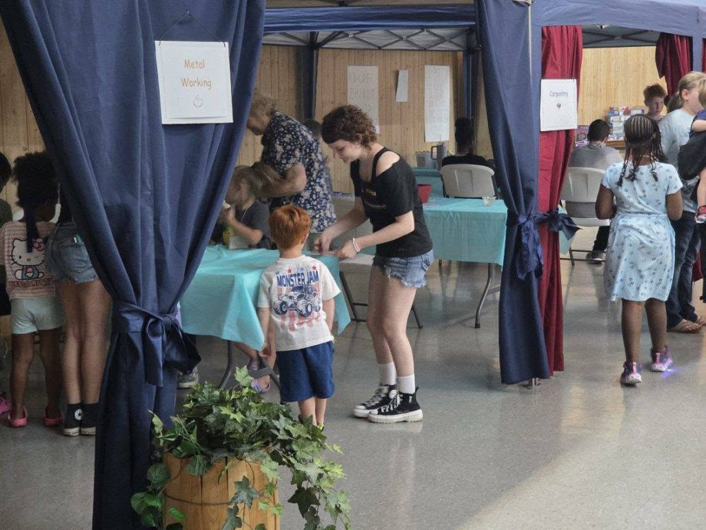 Children and adults participate in indoor workshop activities labeled “Metal Working” and “Carpentry,” with tables, blue curtains, and a potted plant in the foreground.