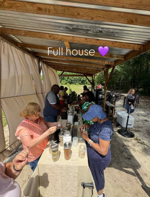 People gathered under a wooden canopy, participating in an outdoor activity with jars and ingredients on tables. Text reads "Full house" with a purple heart emoji.