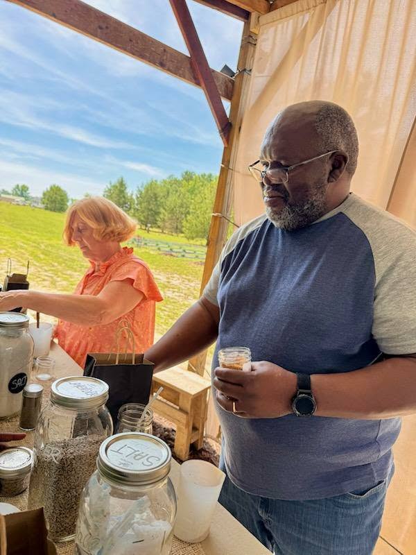 Two people standing at a table with various jars and bags, inside a wooden structure, with a green field and trees visible outside under a blue sky.
