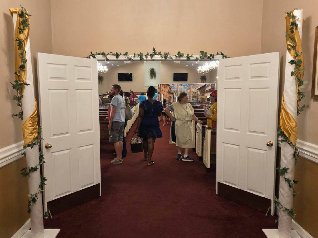 People standing and talking inside a church decorated with white doors, golden ribbon, and greenery, with pews and a mural visible in the background.