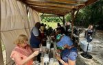 People gathered under a wooden shelter, examining jars on a table, participating in an outdoor workshop or event. Trees and nature are visible in the background.