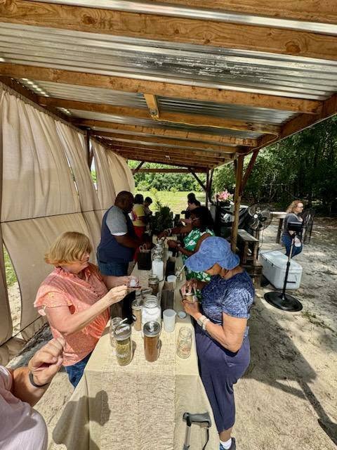 People gathered under a wooden shelter, examining jars on a table, participating in an outdoor workshop or event. Trees and nature are visible in the background.
