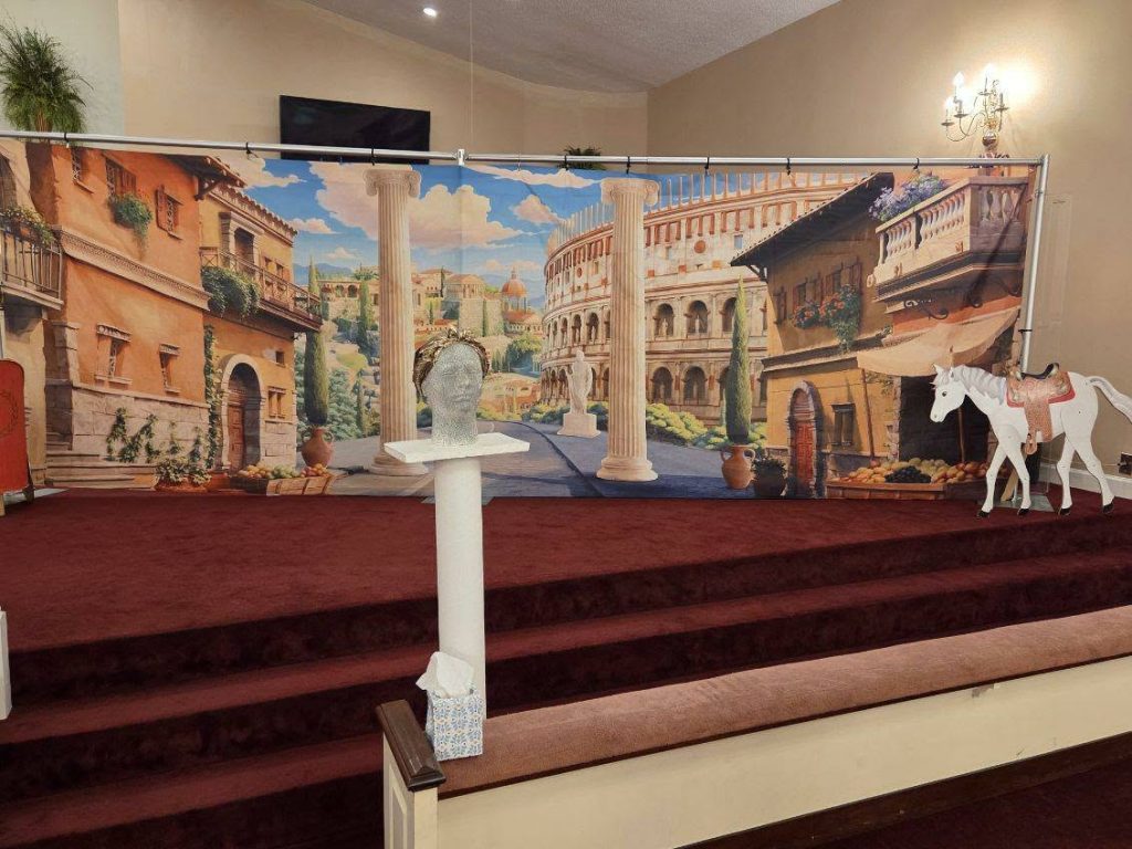 Stage decorated with a Roman city backdrop, fake column, mannequin head with a laurel wreath, and a white cardboard horse with a saddle.
