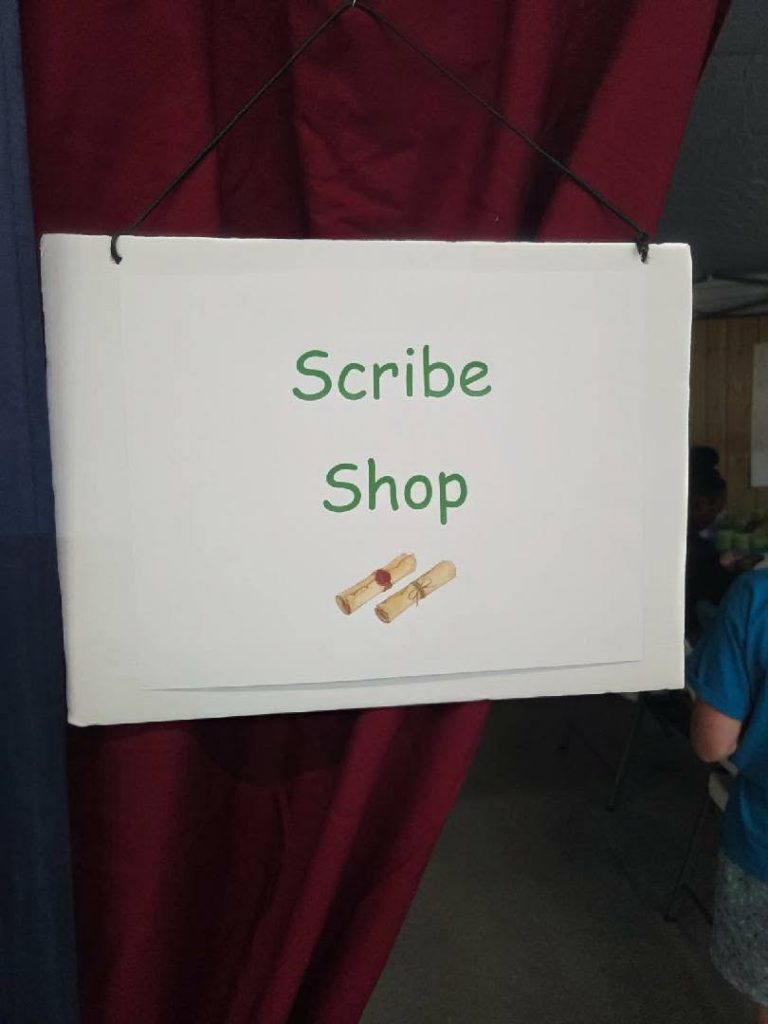 A sign with the words "Scribe Shop" and an illustration of two rolled scrolls, hanging against maroon curtains.