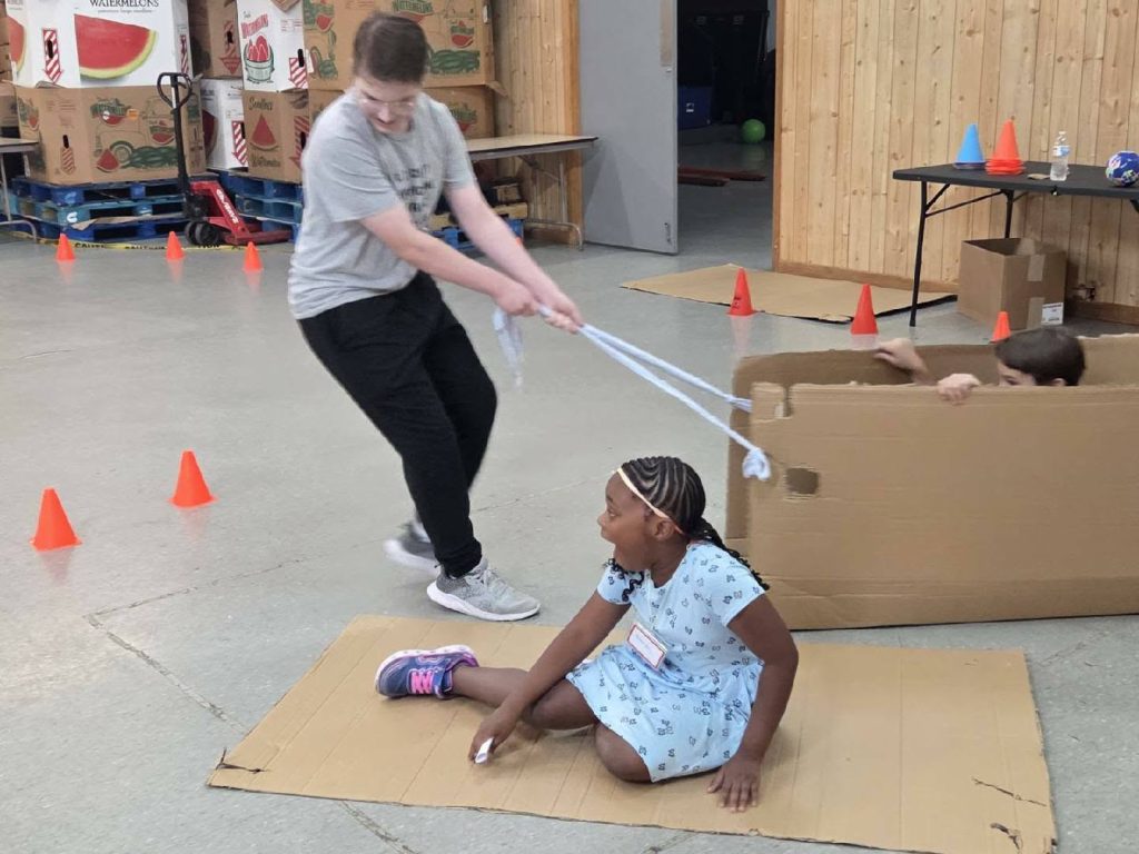 Three children play indoors with cardboard boxes and cones, one girl sits on a flattened box while another child pulls her with a rope.