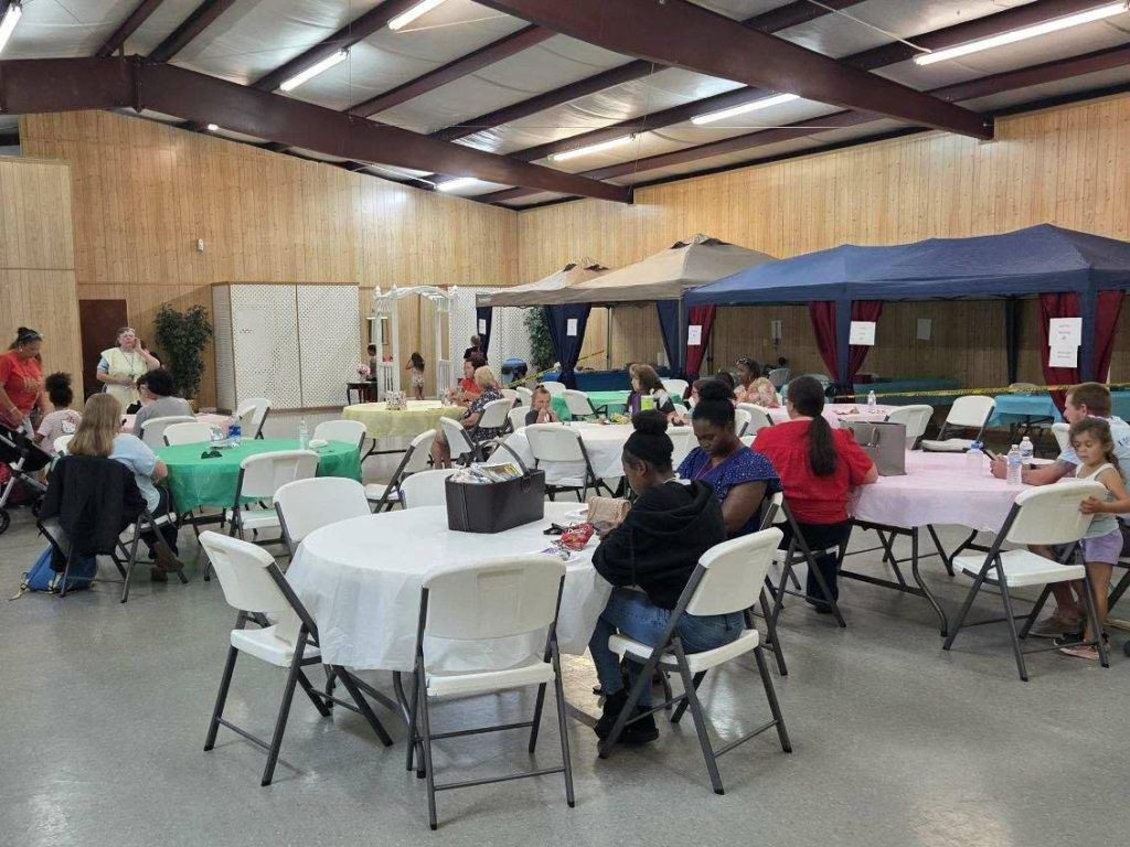 People sit at round tables with colored tablecloths in a large indoor event space, featuring booths with canopies along the wooden-paneled wall.