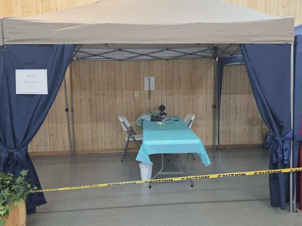 A metal working station under a canopy, with blue curtains, a table and chairs, tools on the table, and caution tape blocking entry.
