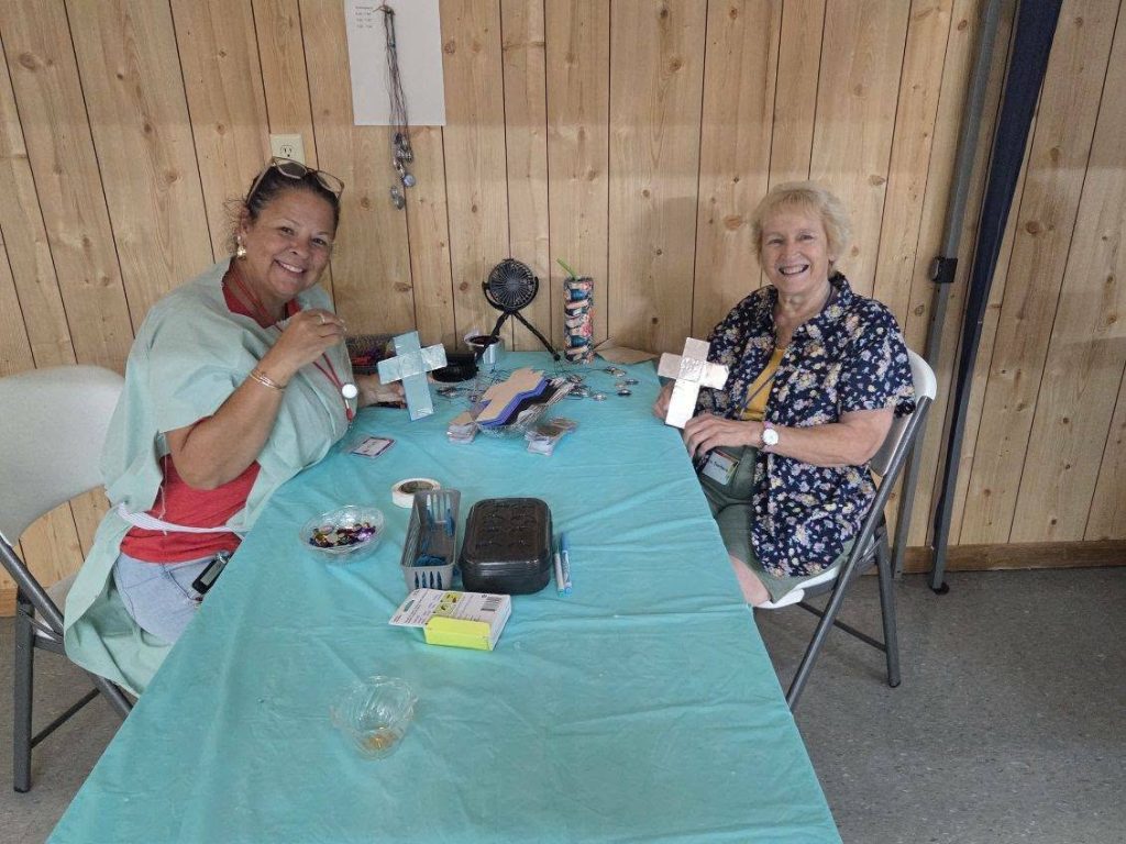Two women sitting at a table with craft supplies, smiling and holding decorated crosses, inside a wooden-paneled room.