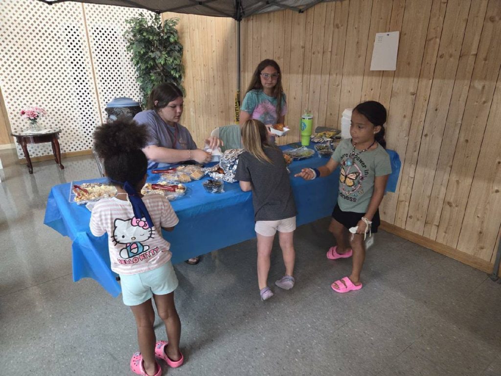 Children and an adult gather at a blue table with snacks in a room with wood-paneled walls, appearing to participate in a bake sale or snack event.