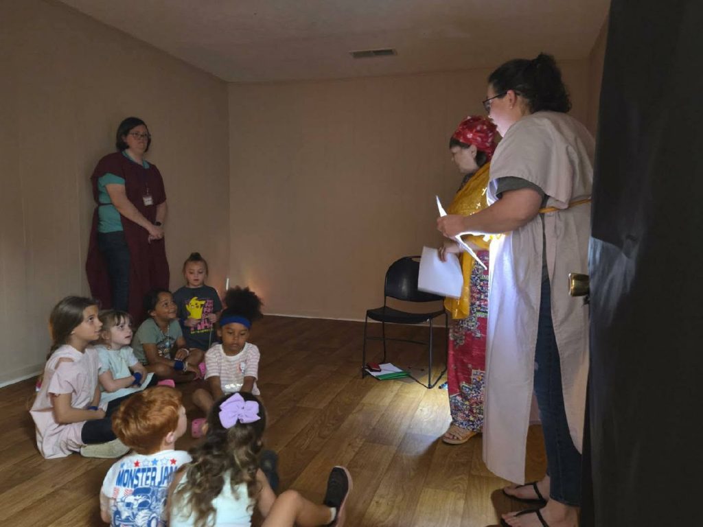 A group of children sit on a wooden floor, listening to two adults in costume and holding papers, while another adult stands nearby against the wall.