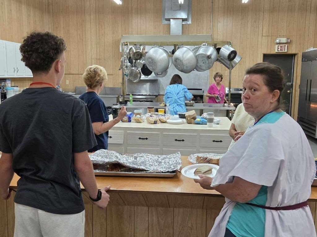 Several people prepare and serve food in a kitchen, with one woman holding a plate and looking toward the camera.