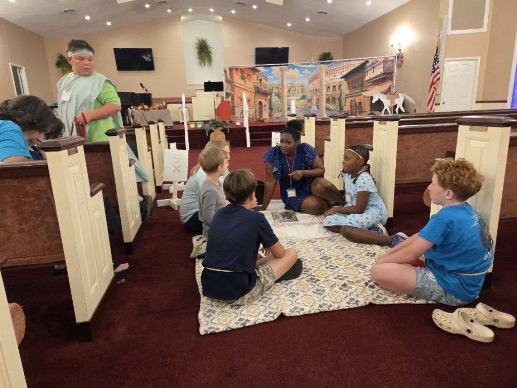 A group of children and an adult sit on a blanket inside a church, engaged in conversation, with colorful decorations and pews around them.
