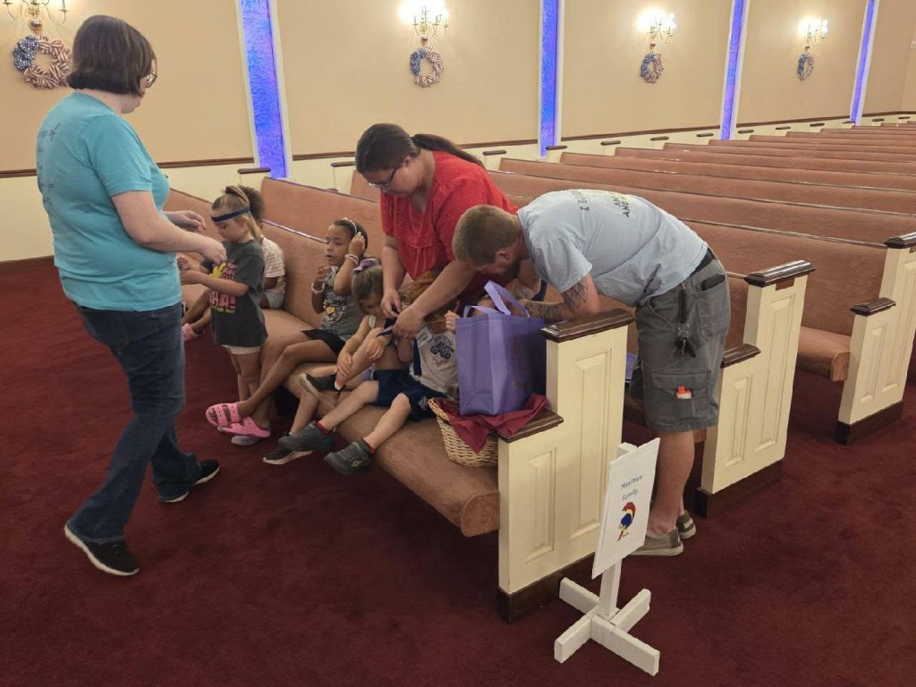 Several adults and children sit and stand in church pews, participating in an activity together. A sign labeled “Nursery Family” stands nearby.