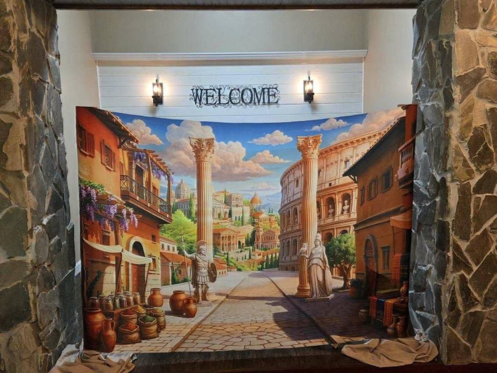 Colorful Roman street backdrop with ancient columns, statues, pottery, and buildings, set under a "WELCOME" sign and two wall lights, between stone pillars.