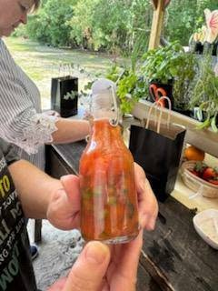 A person holds a small glass bottle filled with homemade red sauce or dressing. Plants, bags, and a bowl are on a rustic wooden table outdoors.