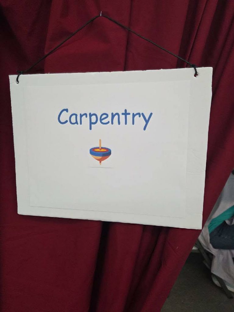 A white sign with the word "Carpentry" and an illustration of a wood lathe, hanging by a string against a red curtain.