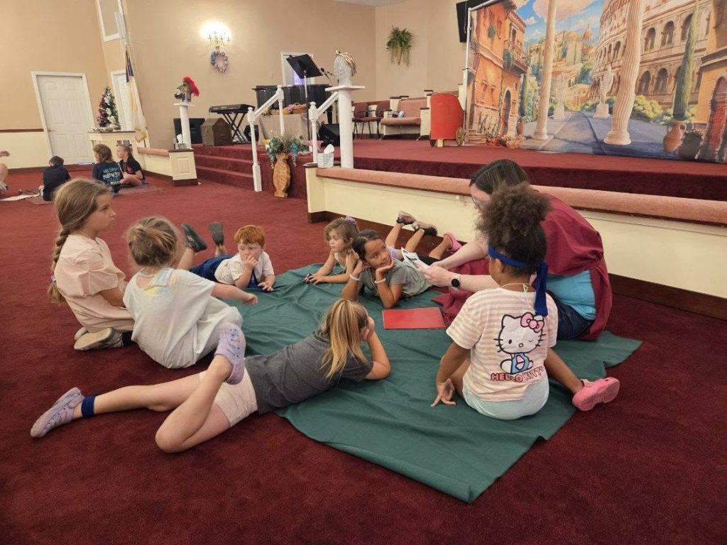 A group of children sits on a green mat in a church hall, listening to an adult reading to them. Other children are sitting in groups nearby.