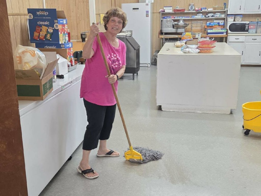 Woman mopping the floor in a kitchen area with snacks and supplies in the background, wearing a pink shirt, black pants, and sandals.