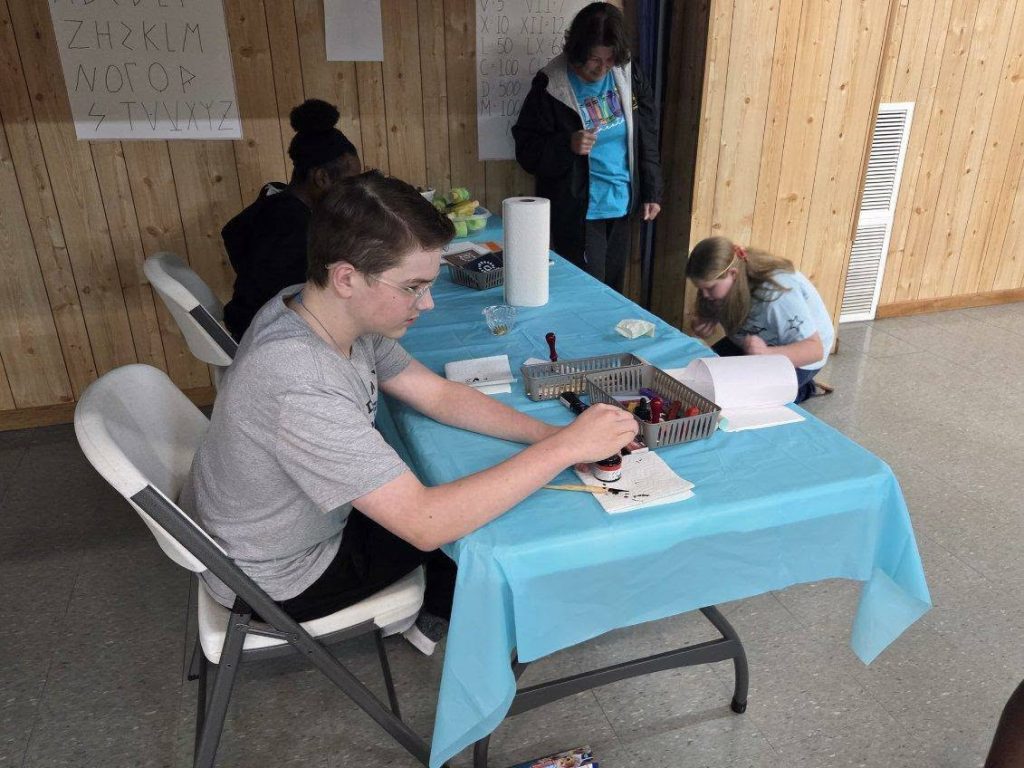 Several young people work at a table covered with a blue cloth, stamping papers, with posters and supplies visible in a wood-paneled room.